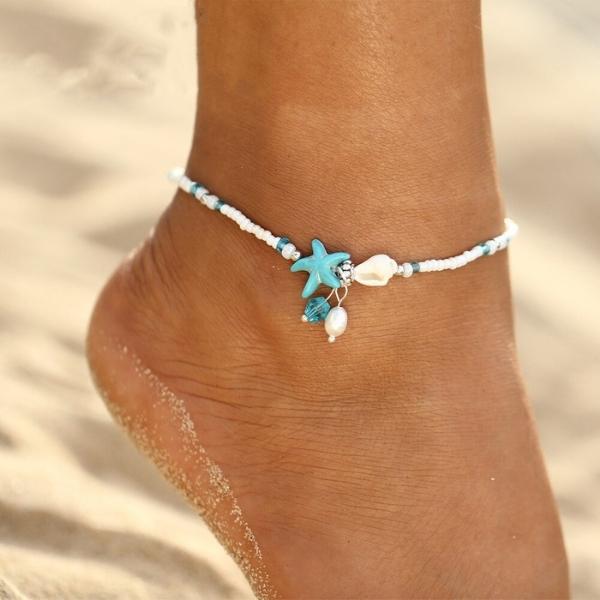 Beach anklet with blue starfish, white beads, and seashell charm on a sandy foot. Perfect summer jewelry accessory for beach lovers.