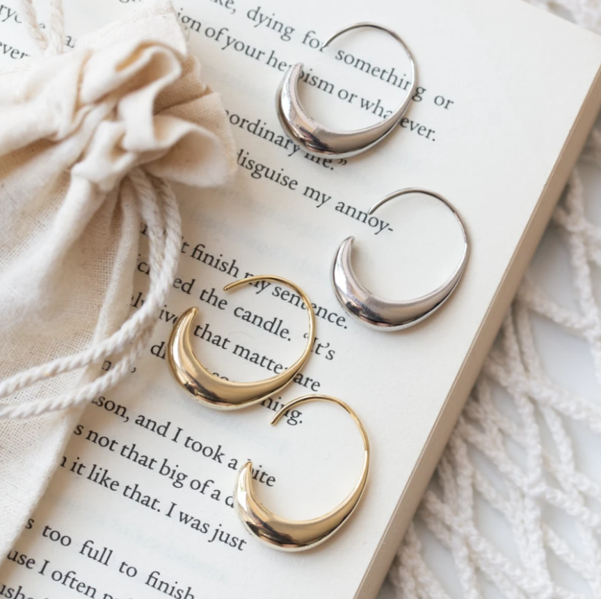 Silver and gold crescent hoop earrings on open book with beige drawstring pouch, stylish jewelry, minimalist fashion accessories, elegant design.