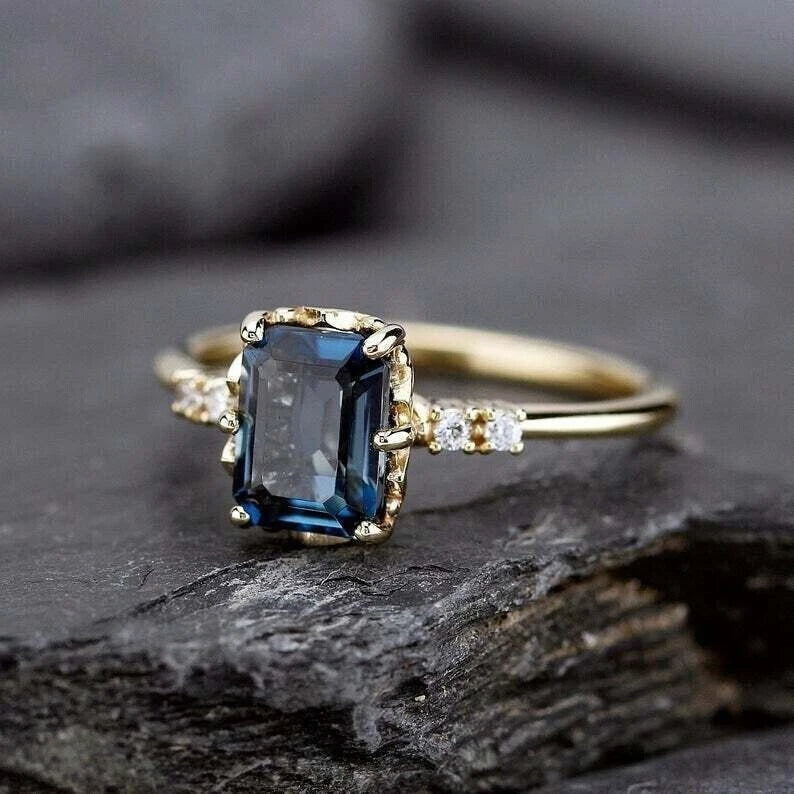 Elegant gold ring with a rectangular blue sapphire centerpiece, accented by small diamonds, displayed on a textured dark stone surface.