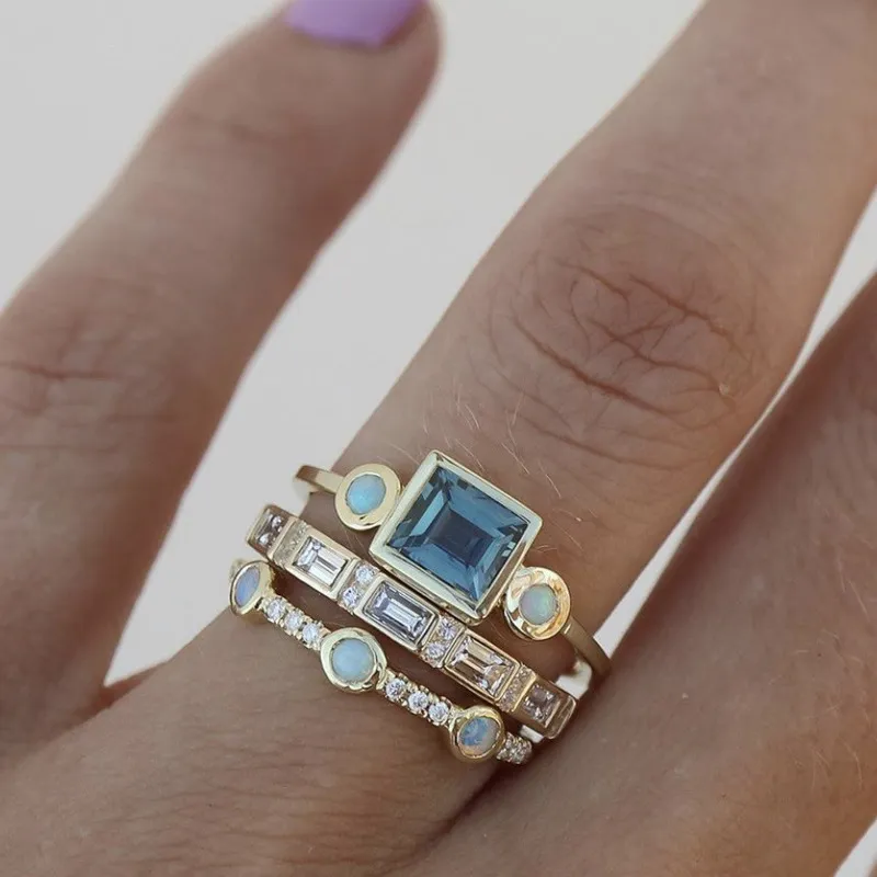 Close-up of a hand wearing a stack of gold rings featuring a large blue gemstone, opals, and diamonds, showcasing elegant jewelry design.