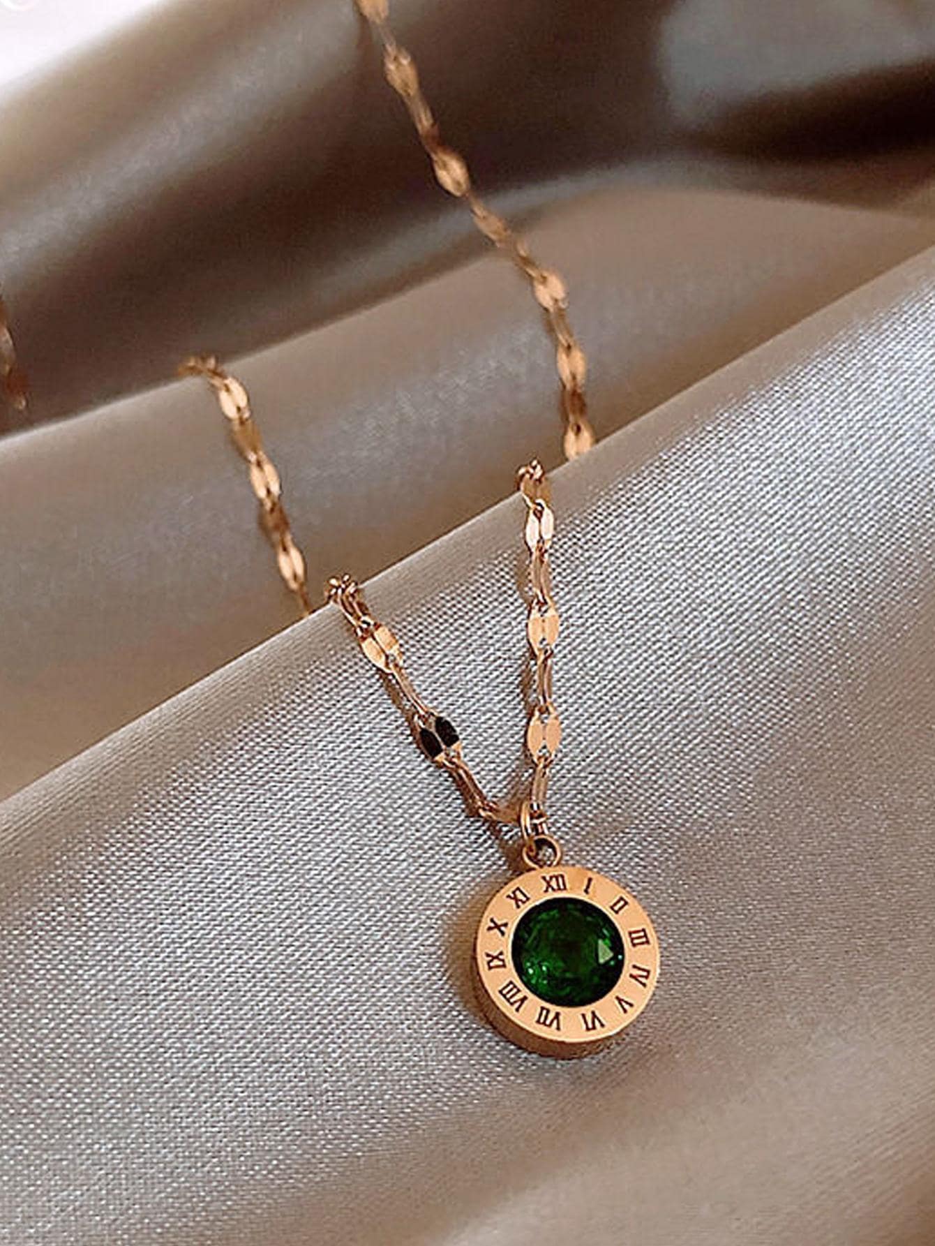 Gold necklace with a round pendant featuring a green gemstone and Roman numerals, elegantly displayed on silky fabric. Luxury jewelry accessory.