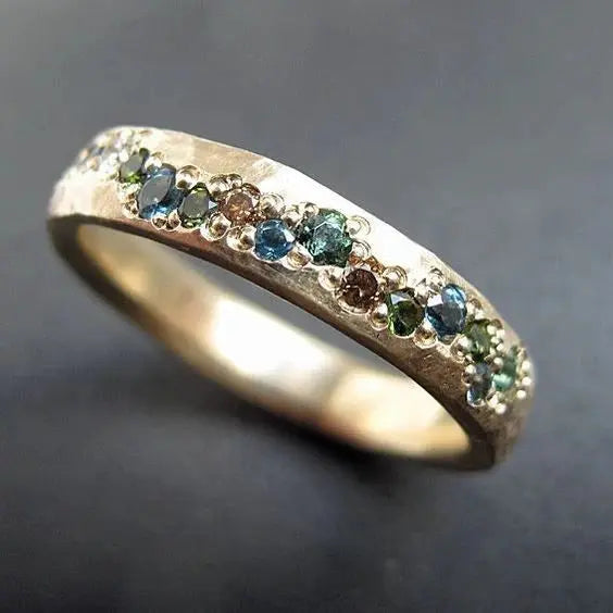 Gold ring with multicolored gemstones, featuring blue, green, and brown stones, set in a textured band. Elegant jewelry design on a dark background.