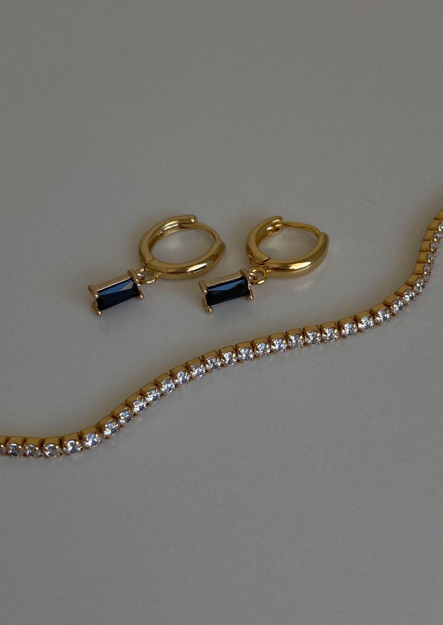 Gold hoop earrings with rectangular blue gemstones and a gold tennis bracelet featuring sparkling diamonds on a neutral background.