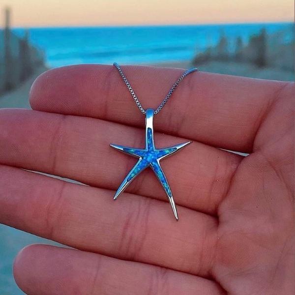 Hand holding a blue opal starfish pendant necklace against a beach background, showcasing ocean-inspired jewelry, perfect for seaside fashion.