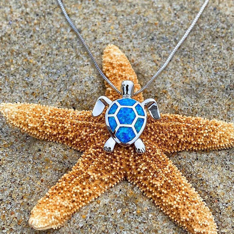 Turtle pendant necklace with blue opal inlay on a starfish, beach sand background. Ocean-themed jewelry, sea turtle charm, coastal fashion accessory.