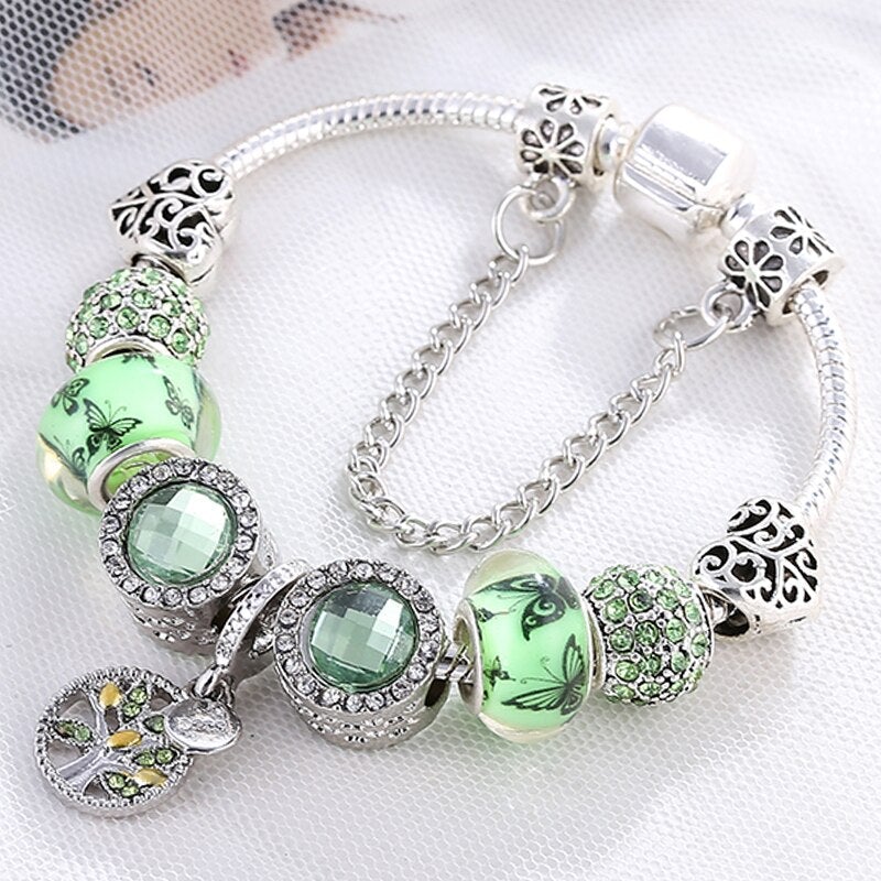 Elegant silver charm bracelet with green crystal beads, floral designs, and a tree of life pendant on a delicate chain. Fashion jewelry accessory.