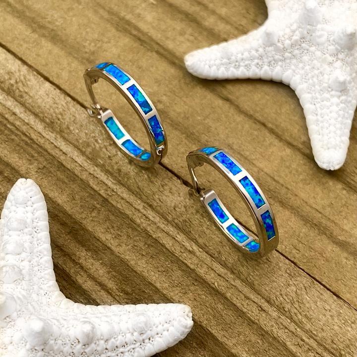Silver hoop earrings with blue opal inlay on wooden surface, surrounded by white starfish. Elegant beach-themed jewelry accessory.