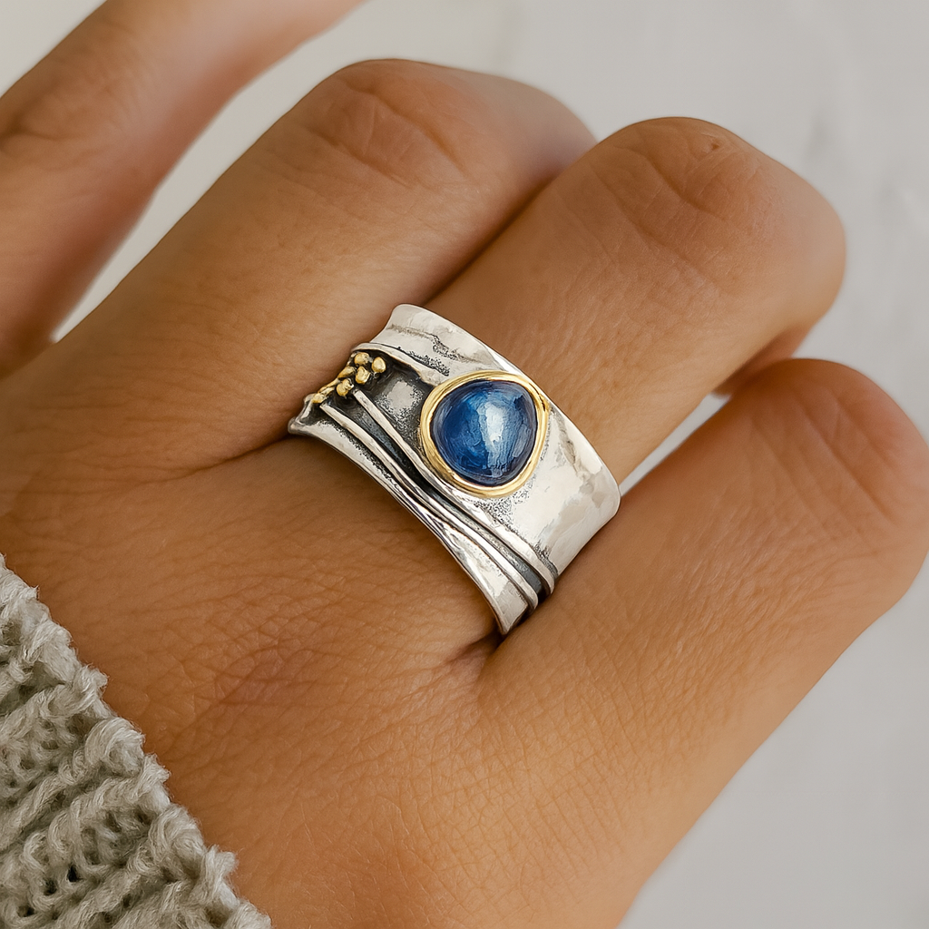 Silver and gold wide band ring with a blue gemstone on a finger, showcasing unique artisan design. Elegant jewelry, handcrafted ring, fashion accessory.
