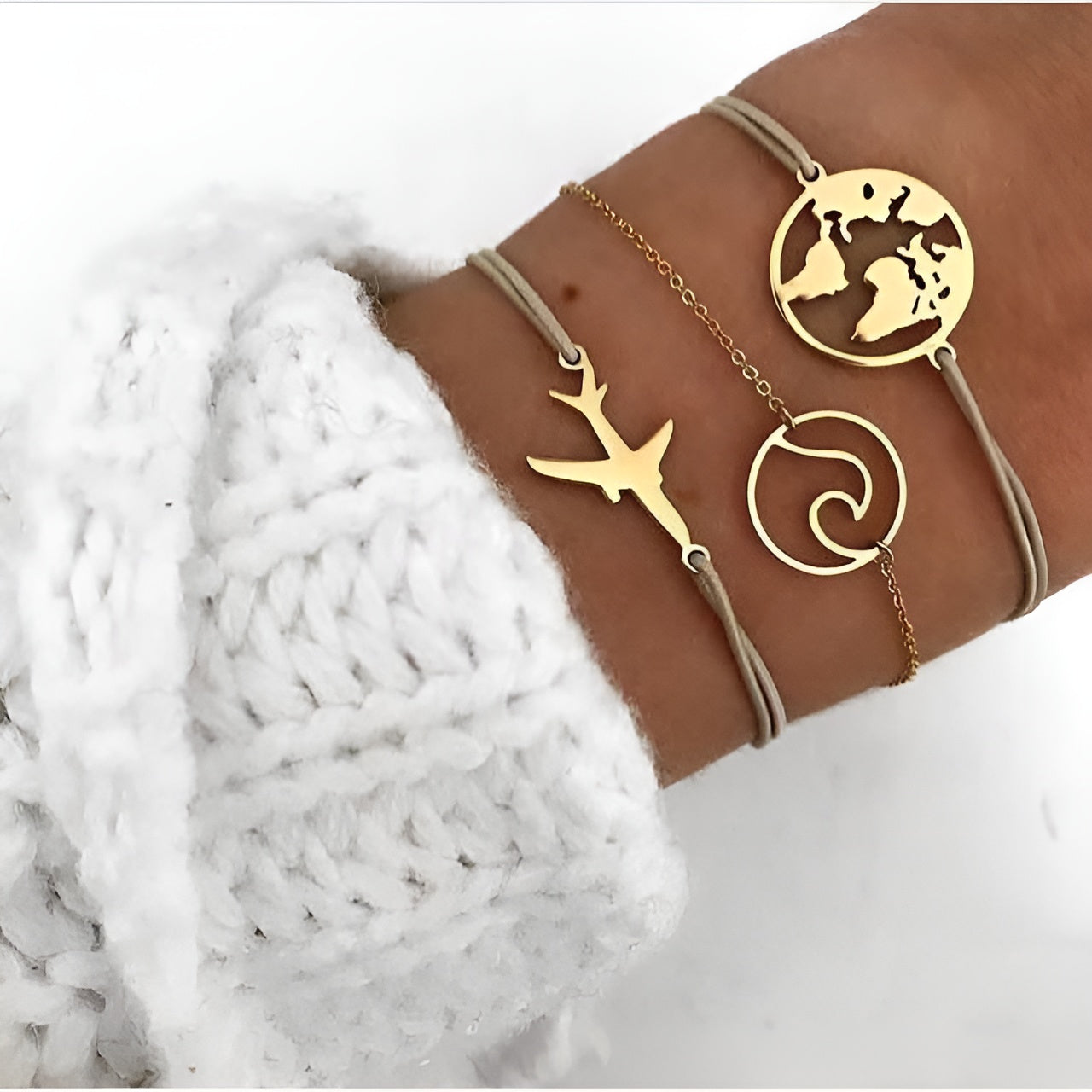 Three gold travel-themed bracelets on wrist: airplane, world map, wave designs. Worn with white knit sweater. Fashion accessories, travel jewelry.