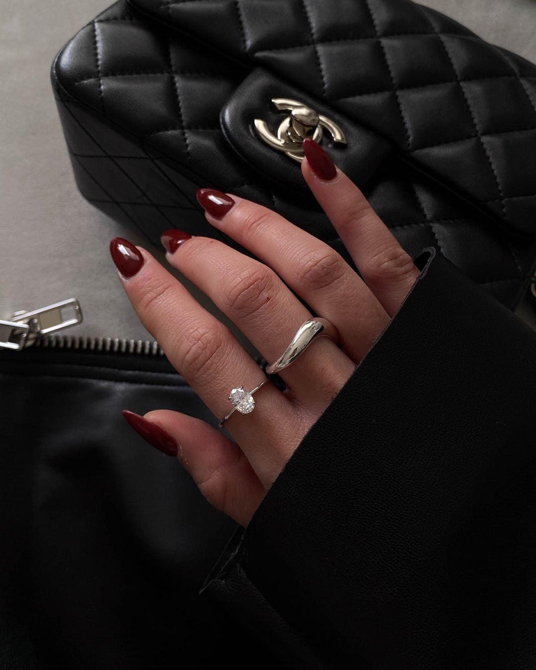 Close-up of a hand with dark red nails wearing a diamond ring and silver band, holding a black quilted designer handbag with a metal logo clasp.