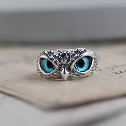 Silver owl ring with intricate detailing and striking blue gemstone eyes, resting on a textured surface. Unique jewelry design, animal-inspired accessory.