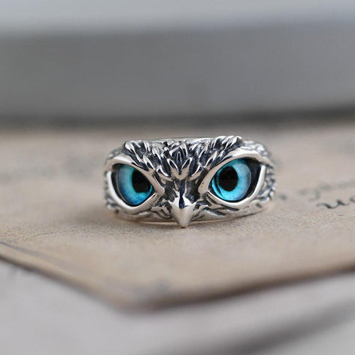 Silver owl ring with intricate detailing and striking blue gemstone eyes, resting on a textured surface. Unique jewelry design, animal-inspired accessory.