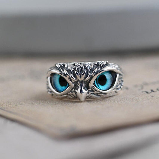 Silver owl ring with intricate detailing and striking blue gemstone eyes, resting on a textured surface. Unique jewelry design, animal-inspired accessory.