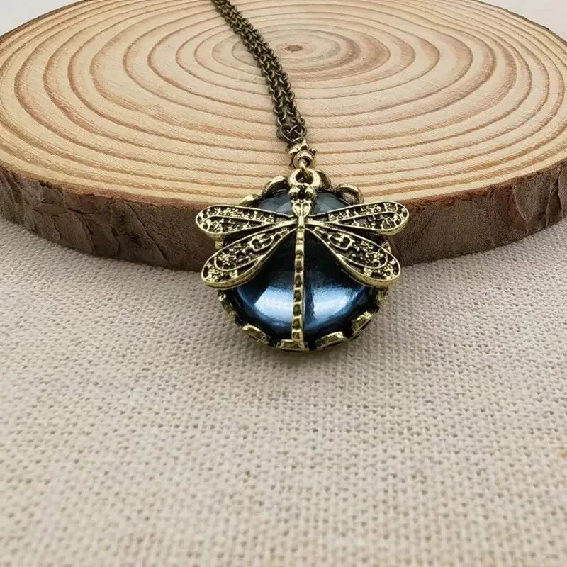 Antique bronze dragonfly pendant necklace on a wooden slice, featuring intricate detailing and a blue gemstone, perfect for vintage jewelry lovers.