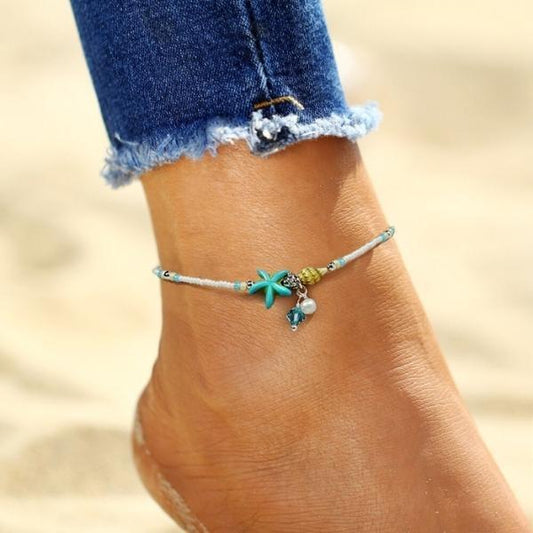 Anklet with starfish charm on foot, worn with frayed jeans on sandy beach. Boho beach jewelry, summer fashion accessory, ocean-themed anklet.