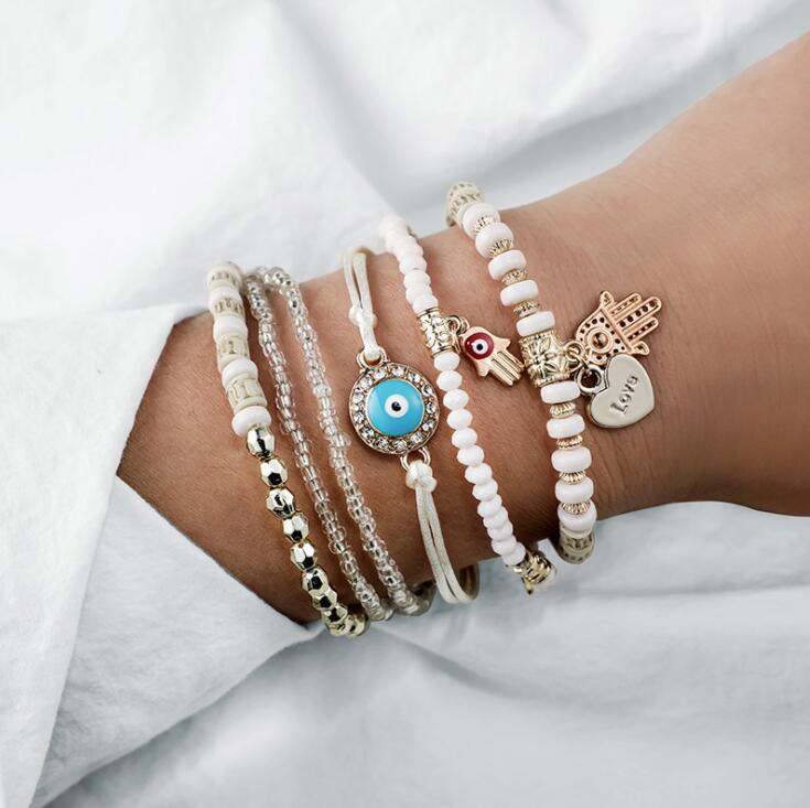 A wrist adorned with multiple bohemian-style bracelets featuring beads, an evil eye charm, and a heart pendant, showcasing trendy fashion accessories.