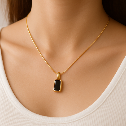 Gold necklace with a rectangular black pendant on a woman's neck, wearing a white top. Elegant jewelry, minimalist style, fashion accessory.