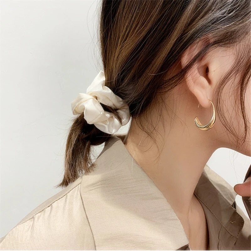 Woman with brown hair in a low ponytail, wearing a white scrunchie and gold hoop earrings, beige shirt, side profile. Fashion accessories.