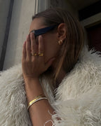Person wearing sunglasses, gold rings, and bracelets with a white fur coat.