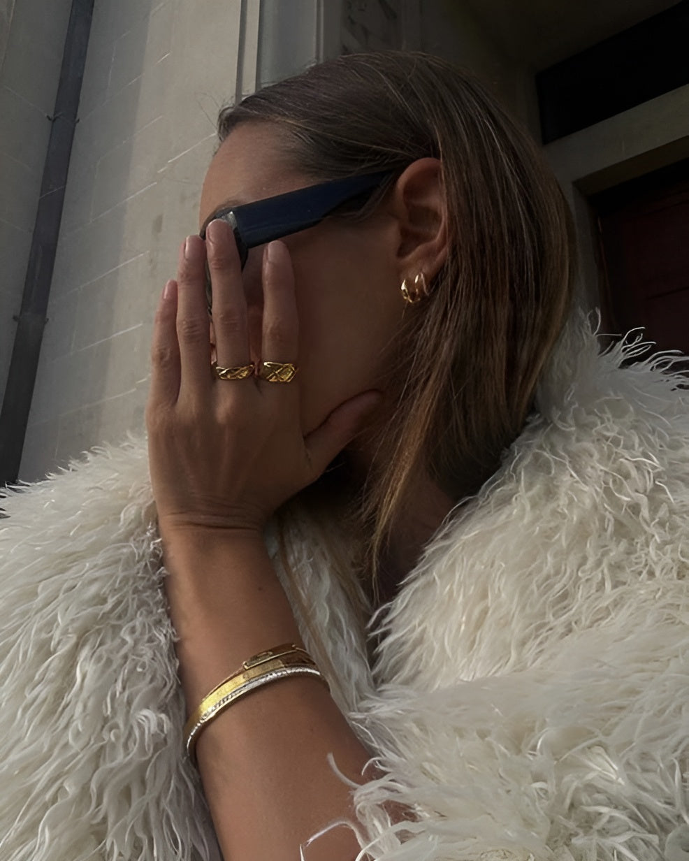 Person wearing sunglasses, gold rings, and bracelets with a white fur coat.
