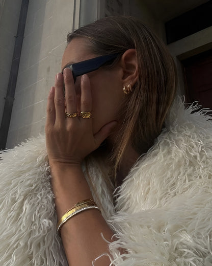 Person wearing sunglasses, gold rings, and bracelets with a white fur coat.