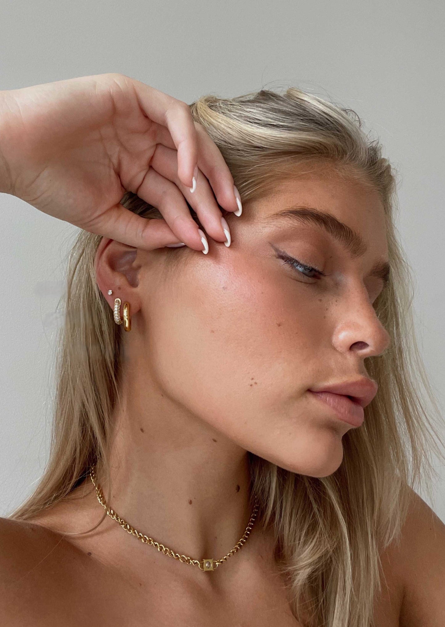 Close-up of a woman with blonde hair and gold jewelry against a neutral background