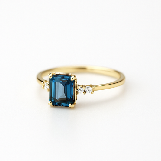 Gold ring with an emerald-cut blue sapphire centerpiece, flanked by small diamonds, on a white background. Elegant jewelry, luxury accessory.