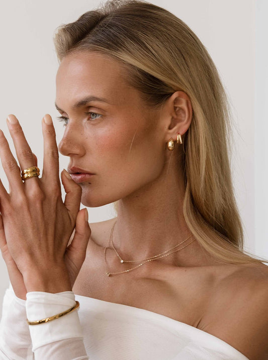 Woman wearing gold jewelry, including hoop earrings, layered necklaces, rings, and a bracelet, with a white off-shoulder top, showcasing elegant fashion accessories.