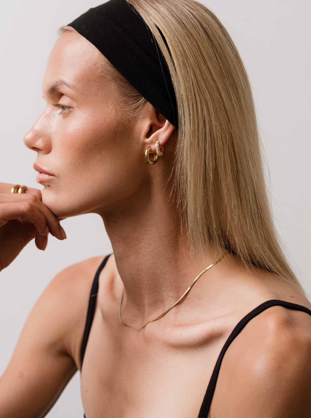 Profile of a woman with long blonde hair wearing a black headband, gold hoop earrings, and a thin necklace, showcasing minimalist fashion style.