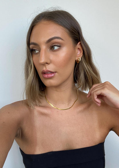Young woman with shoulder-length hair, wearing gold hoop earrings and a necklace, in a black strapless top, looking to the side against a white background.