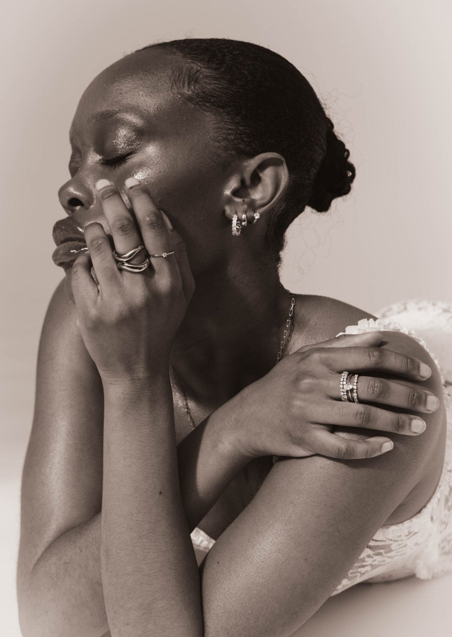 Elegant woman with closed eyes, wearing multiple rings and earrings, showcasing skincare and jewelry. Black and white portrait, serene expression.