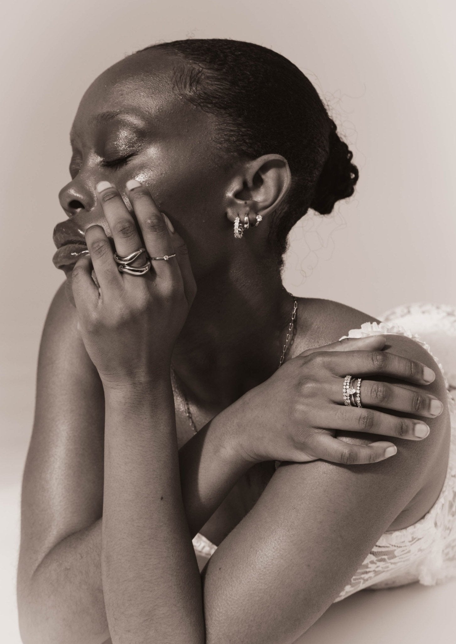 Elegant woman with closed eyes, wearing multiple rings and earrings, showcasing skincare and jewelry. Black and white portrait, serene expression.