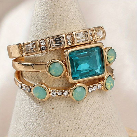 Gold ring set with turquoise and clear gemstones, featuring a large rectangular blue stone, displayed on a beige fabric cone. Elegant jewelry accessory.