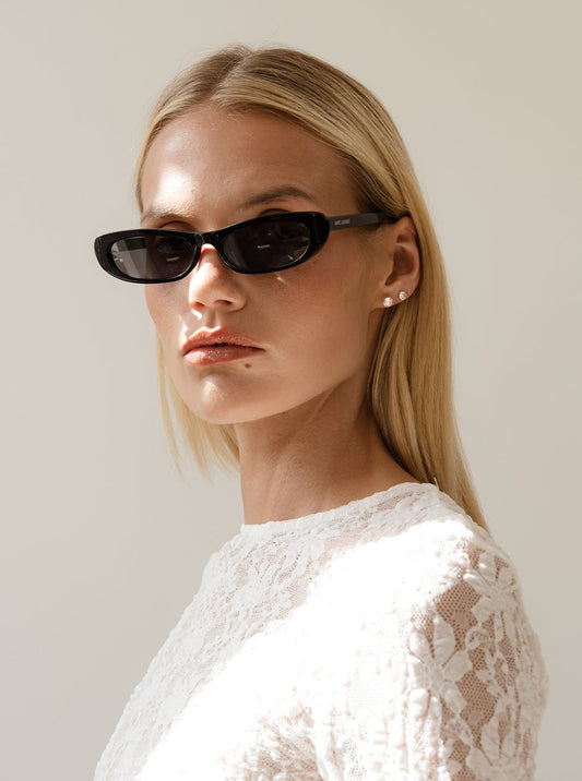 Blonde woman wearing black oval sunglasses and a white lace top, posing against a neutral background. Fashionable eyewear, chic style.