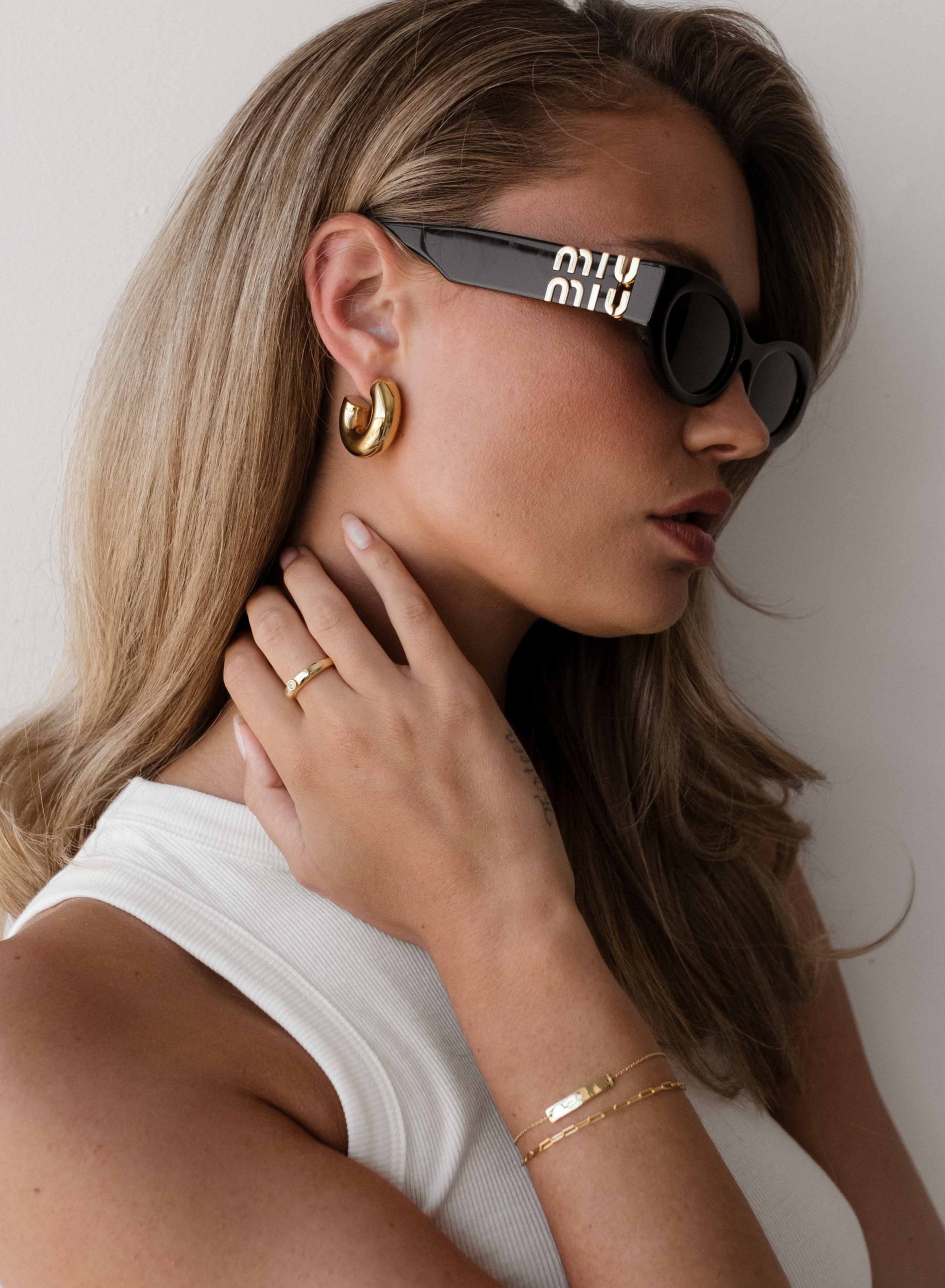 Woman wearing black sunglasses with a visible brand logo, gold earrings, ring, and bracelet against a neutral background.
