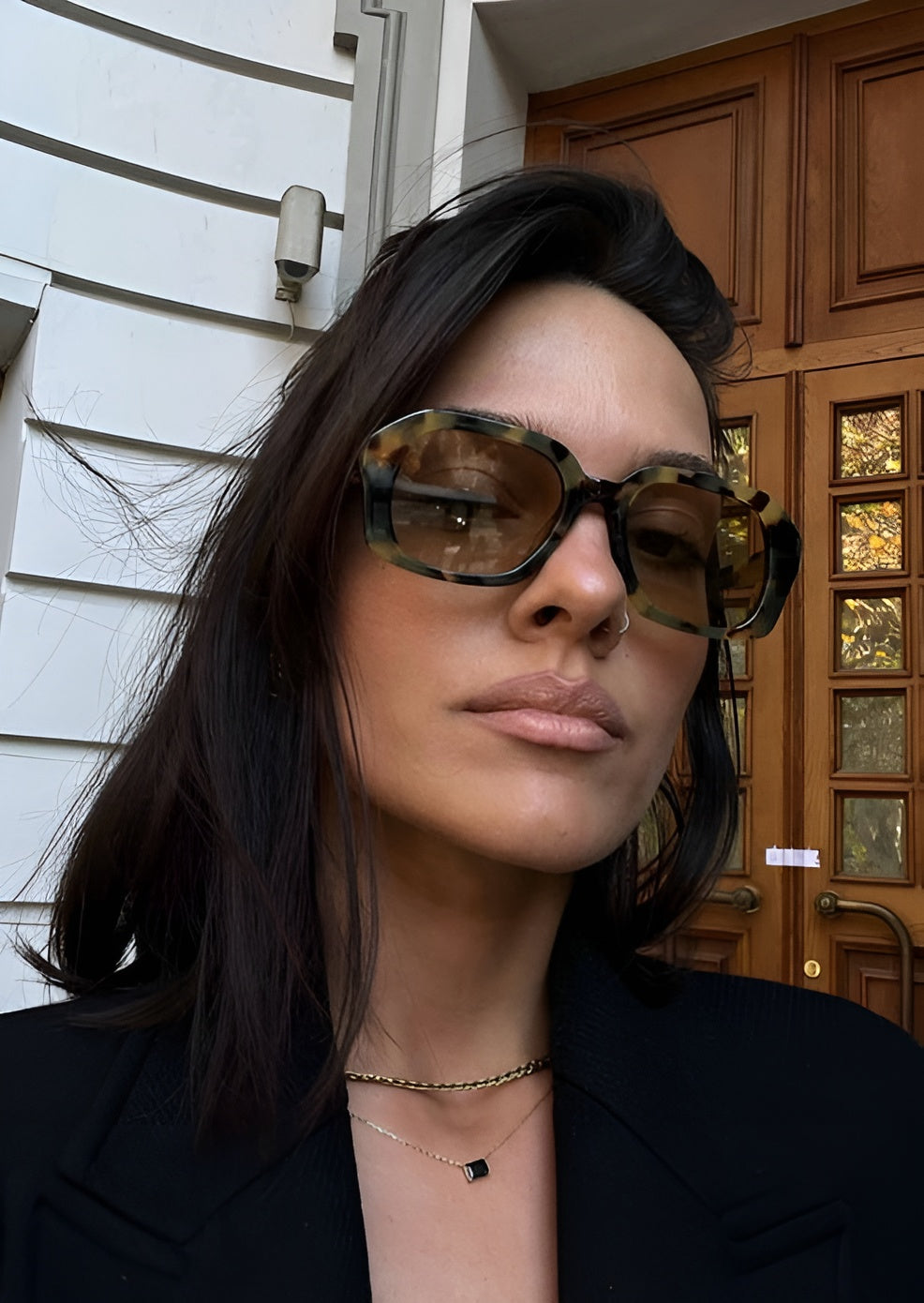 Woman wearing sunglasses with a neutral expression, standing in front of a wooden door.