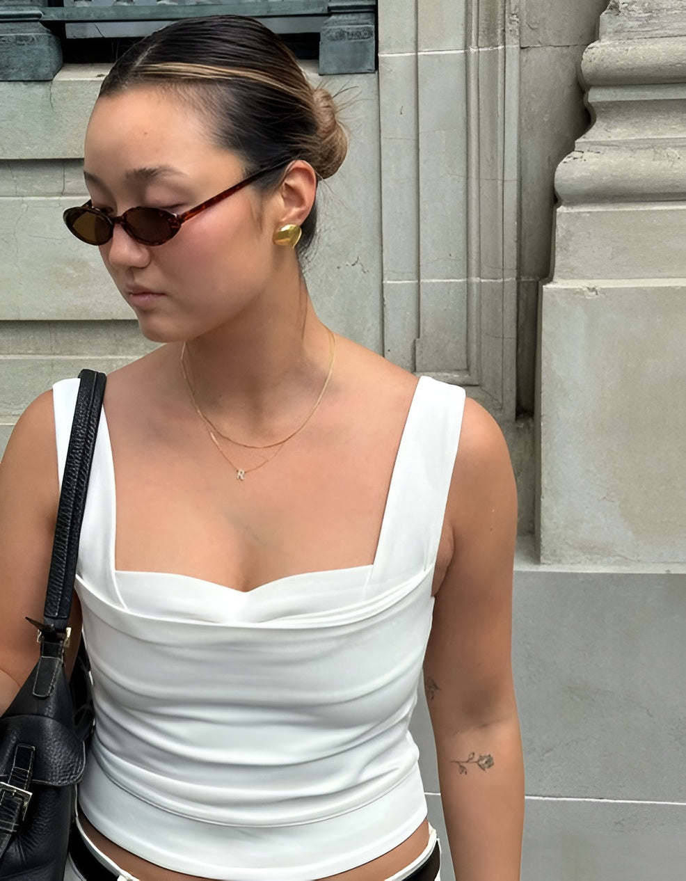 Woman wearing a white top and sunglasses, standing against a stone wall.