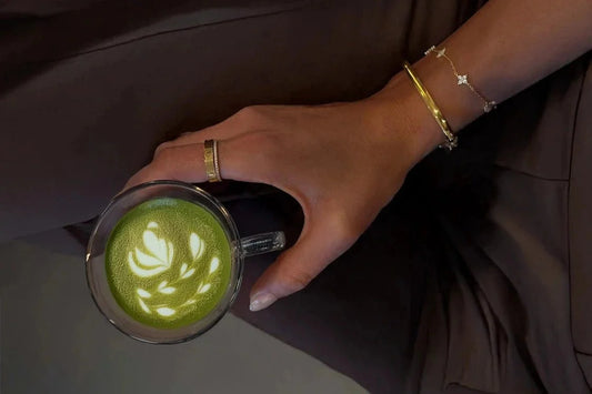 Hand holding a cup of green matcha latte with intricate patterns, wearing gold bracelets.