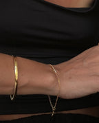 Close-up of a woman's crossed arms wearing a gold bangle and delicate chain bracelet, against a black top. Fashion accessories, jewelry style.