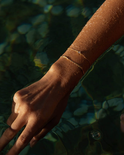 Hand with delicate gold bracelet submerged in shimmering water, highlighting jewelry elegance and summer vibes.