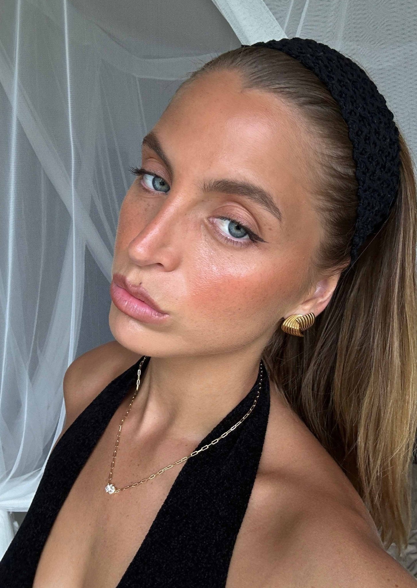 Woman with black headband and halter top, wearing gold earrings and necklace, posing indoors. Fashion, beauty, and accessories.