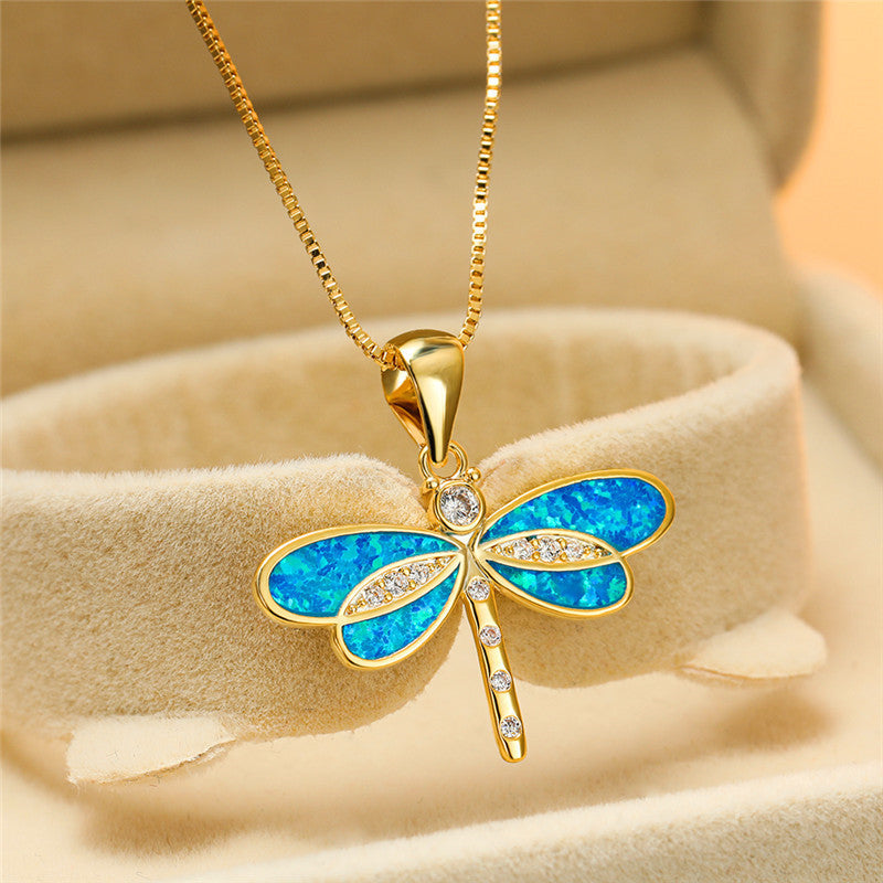 Gold dragonfly necklace with blue opal wings and diamond accents, displayed in a beige jewelry box. Elegant pendant, perfect for gifts.