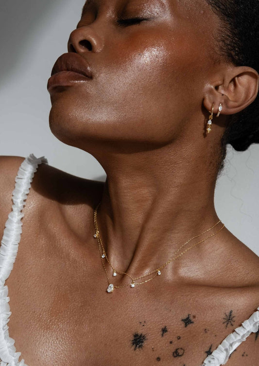 Woman wearing gold layered necklaces and hoop earrings, showcasing elegant jewelry against a tattooed shoulder. Fashion accessories, jewelry trends.