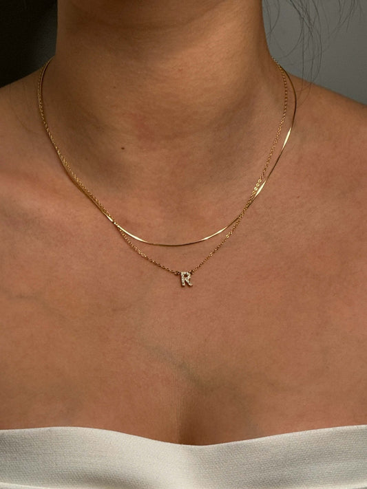Gold layered necklaces on woman's neck, featuring a delicate chain with an "R" pendant. Elegant jewelry, minimalist style, fashion accessory.