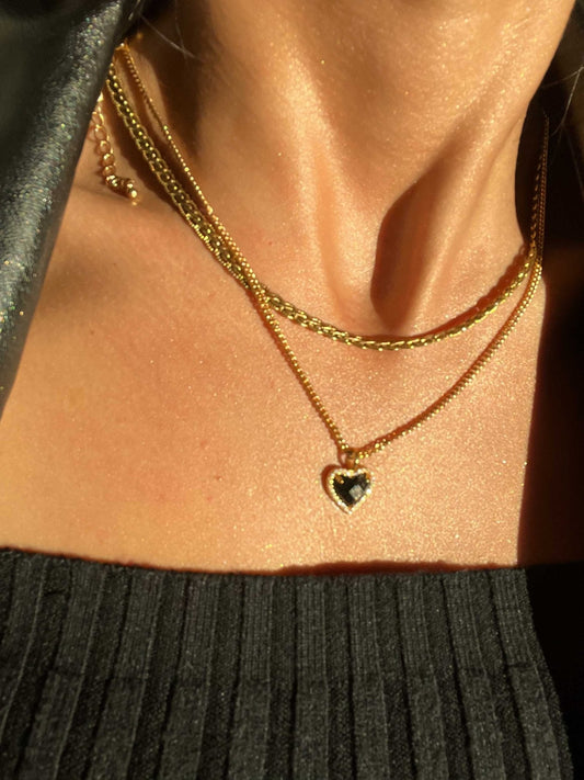 Gold layered necklaces with a heart-shaped pendant on a woman's neck, wearing a black ribbed top. Elegant jewelry fashion, close-up.