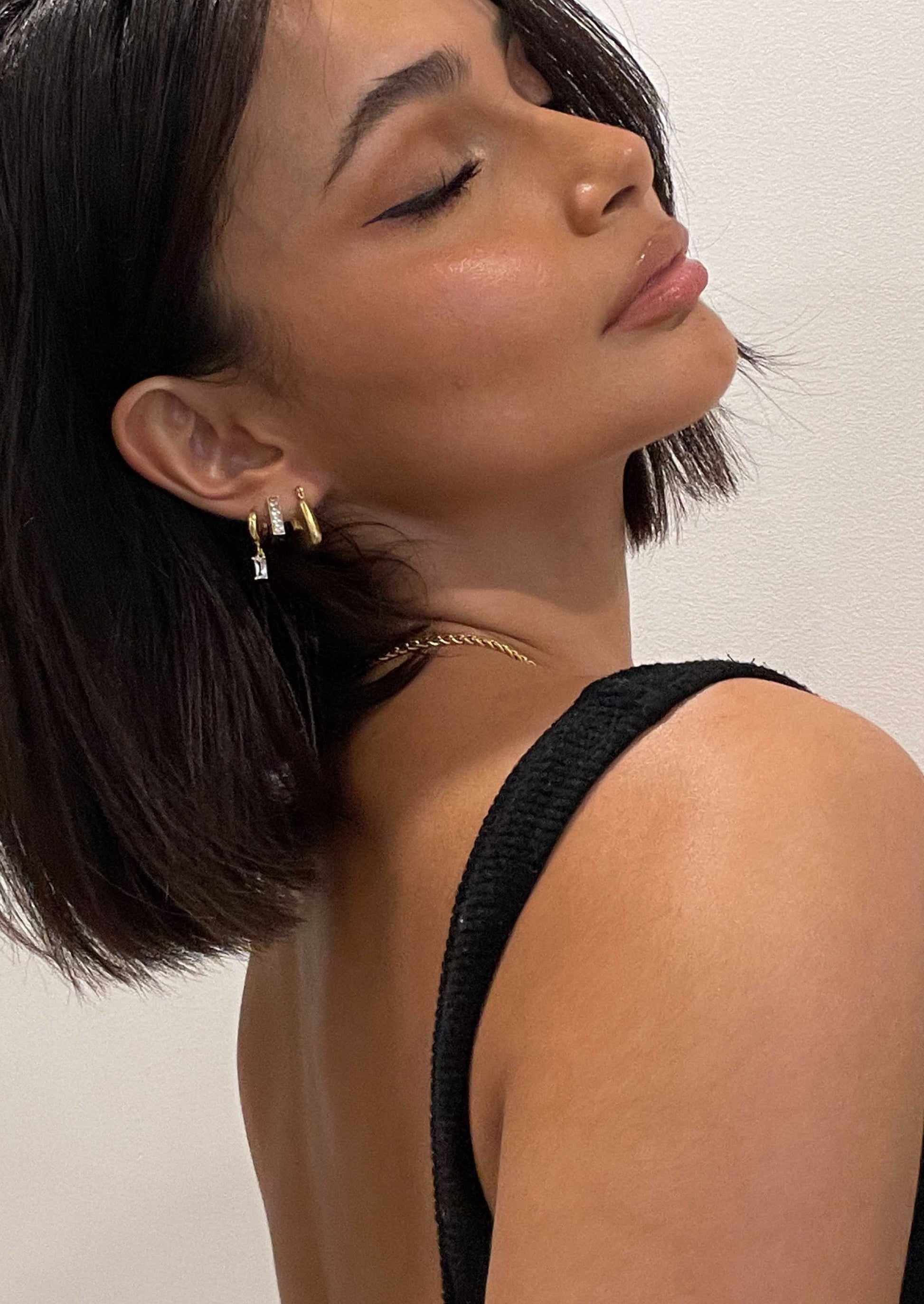 Woman with short dark hair wearing gold hoop earrings and a black top, showcasing a minimalist jewelry style against a plain background.