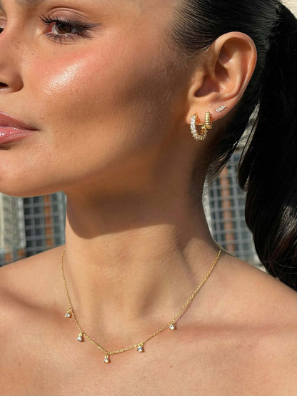 Close-up of a woman wearing gold hoop earrings with diamonds and a delicate gold necklace with small diamond pendants, against a blurred cityscape.