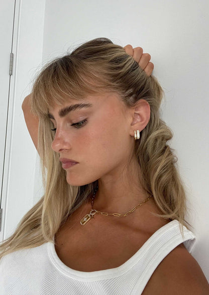 Young woman with blonde hair and bangs, wearing gold hoop earrings and a chain necklace, poses in a white tank top against a white background.