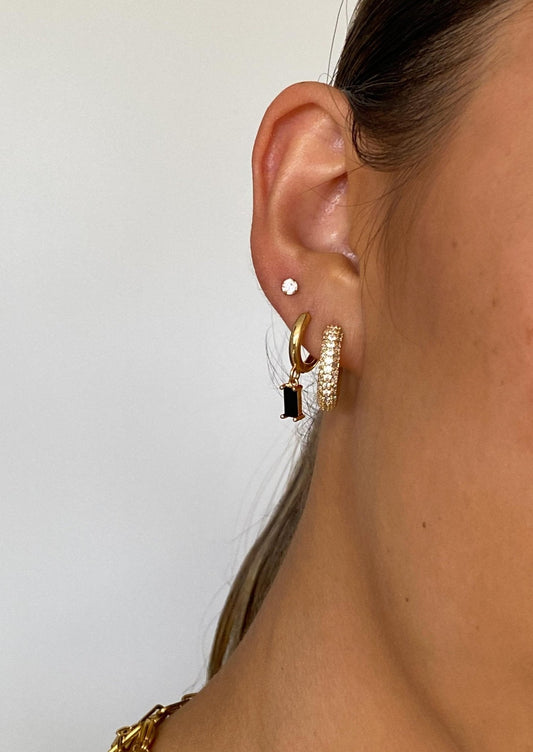 Close-up of a woman's ear with multiple gold hoop earrings, a black gemstone earring, and a small diamond stud, showcasing trendy ear jewelry.