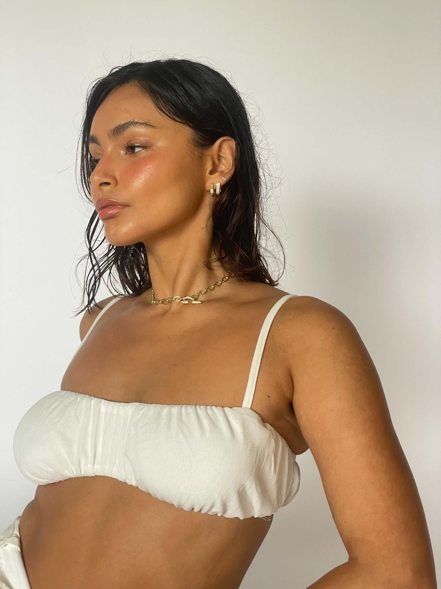 Woman in white bikini top with gold jewelry, wet hair, and natural makeup against a plain background. Fashion, swimwear, summer style.
